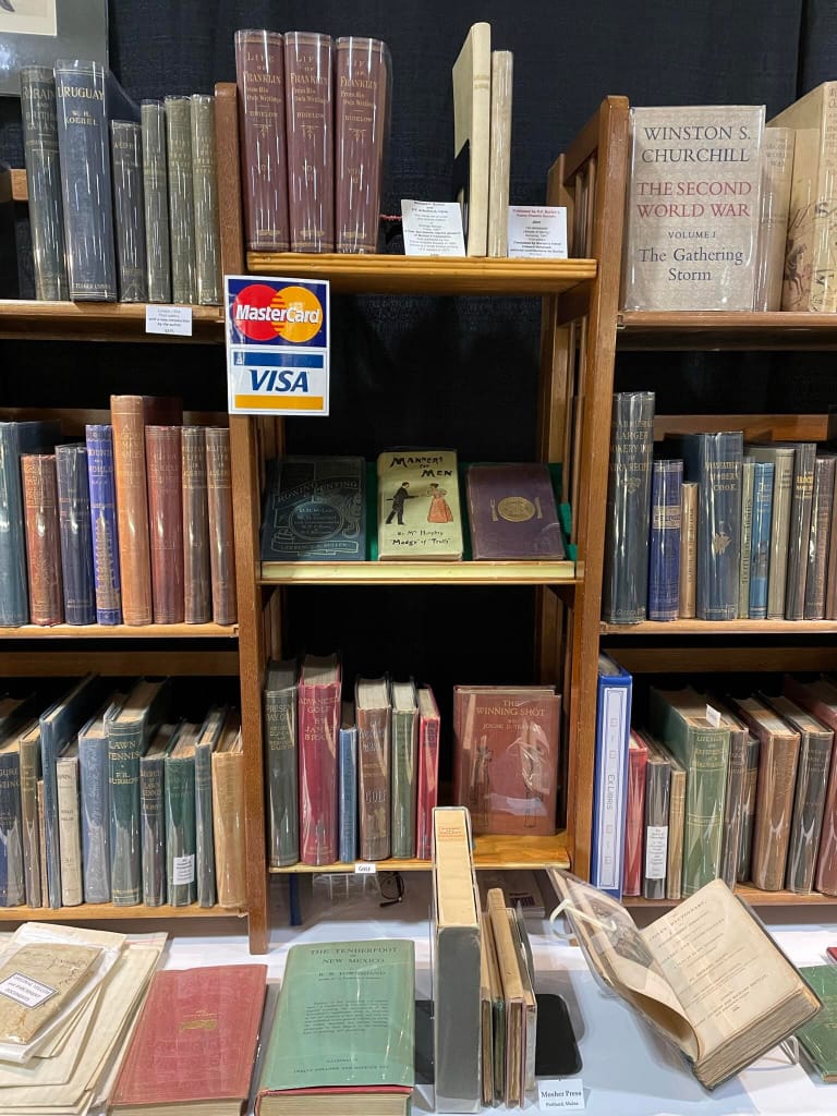 A book fair display of antiquarian books, all of which bear a protective plastic film over their bindings, and one propped open on a desk placed in front of three collapsible wooden shelves displaying the rest